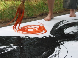 Medium shot photograph of black calligraphy stroke with red stroke painted by workshop participant using a big brush on white paper outside on the grass where you also see the bare feet of the painter